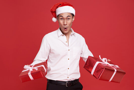 Young asian businessman wearing Santa hat and holding gift box in christmas party on red background. Happy new year concept.