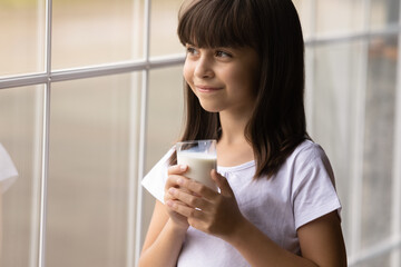 Happy cute thoughtful girl drinking milk, holding glass, looking out of window at home, smiling at good thoughts, thinking. Kid getting calcium, vitamins, probiotics, proteins for growth from dairy