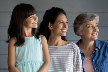 Fototapeta premium Three female generations family head shot portrait. Happy little daughter kid, mother, grandmother standing together in line, looking away, thinking of future vision, smiling at good thoughts