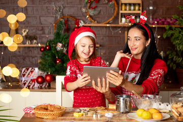 mom and daughter are talking via video link, passing greetings on a tablet in a dark kitchen with a Christmas tree for New Year or Christmas, smiling and having fun together in a Santa Claus hat