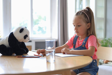 Smilling happy girl sitting in the table with a toy panda bear near to her enjoying creative...