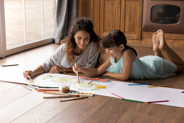Happy Latin daughter kid and mom painting in water colors, lying on heating parquet floor in...
