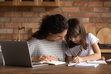 Best mom helping schoolkid daughter girl to do school homework. Mother watching kid writing in copybook at table with laptop, book, textbook. Parent support, education, childhood concept