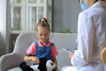 Little girl at the doctor for a checkup. Doctor playfully checking the heart beat of a teddy bear.