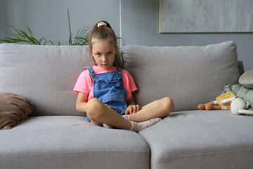 Sad little girl sitting on sofa alone at home.