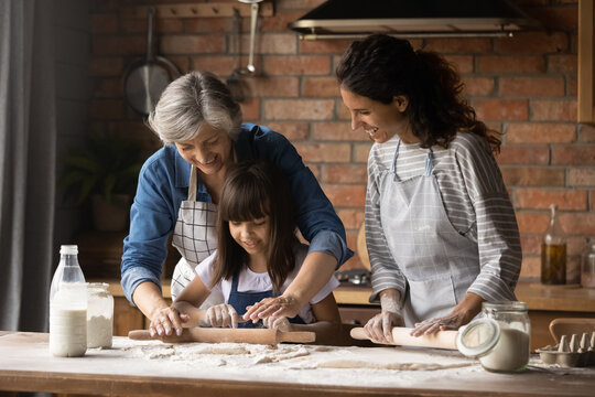 Happy Hispanic mom and grandma teaching girl to bake, kneading, rolling dough for pastry together. Three family female generations cooking homemade bakery food, having fun at messy kitchen table