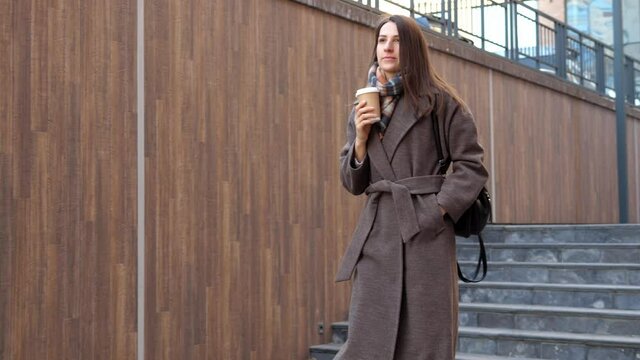 Beautiful young woman is walking in the middle of a modern cityscape in a coat. Happy young woman enjoying life