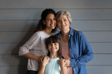 Fototapeta na wymiar Happy girl, young mother, senior grandmother head shot portrait. Kid, mom, grandma standing at grey wall, hugging with love, affection, looking at camera, smiling. Three female family generations