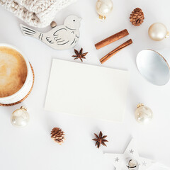 Christmas still life scene. Blank paper greeting card, cinnamon sticks, cup of hot chocolate, nordic decorations, pine cones, knitted hat on white desk table. Hygge, cozy home, Scandinavian aesthetic