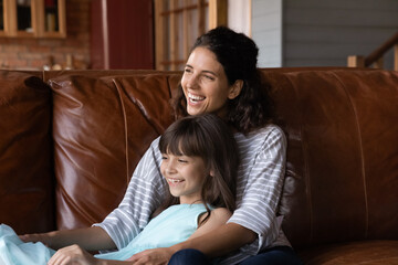 Happy mother holding sweet daughter in arms, relaxing on leather couch at home, looking away,...