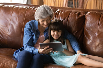 Happy loving older grandma and cheerful granddaughter girl making video call on tablet computer together, relaxing on couch with digital gadget, watching movie, reading on modern tech device
