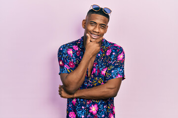 Young black man wearing hawaiian shirt and sunglasses looking confident at the camera smiling with crossed arms and hand raised on chin. thinking positive.