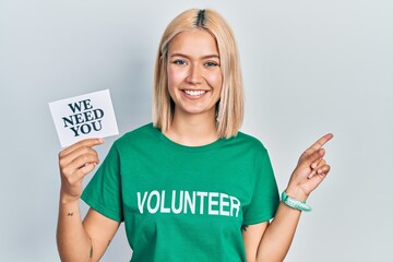 Beautiful blonde woman wearing volunteer t shirt showing we need you banner smiling happy pointing with hand and finger to the side