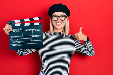 Beautiful blonde woman holding video film clapboard smiling happy and positive, thumb up doing...