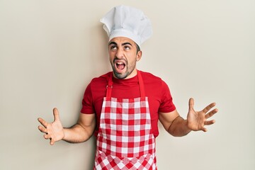 Young hispanic man wearing baker uniform crazy and mad shouting and yelling with aggressive expression and arms raised. frustration concept.