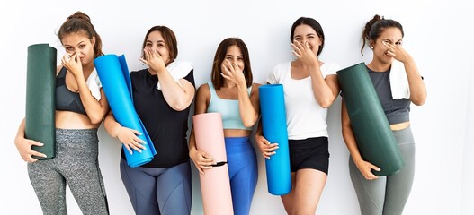 Group of women holding yoga mat standing over isolated background smelling something stinky and...