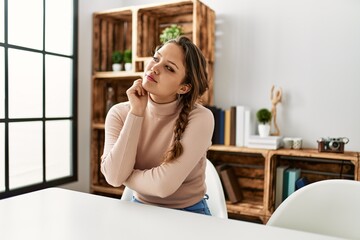 Young hispanic girl wearing casual clothes sitting on the table at home serious face thinking about...