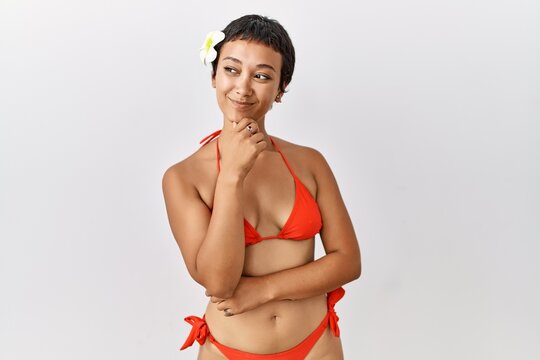 Young hispanic woman with short hair wearing bikini looking confident at the camera smiling with crossed arms and hand raised on chin. thinking positive.