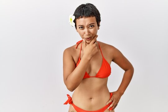 Young hispanic woman with short hair wearing bikini looking fascinated with disbelief, surprise and amazed expression with hands on chin