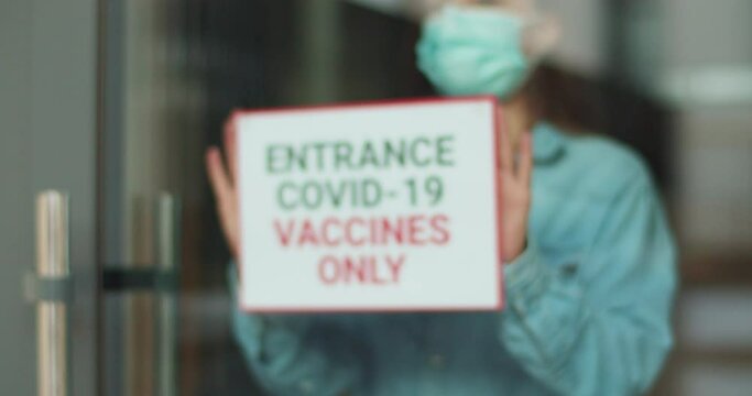 Plate with the inscription Entrance Covid-19 vaccines only on the entrancedoor. Woman wearing face mask attaching paper with text on the glass in pandemic time.