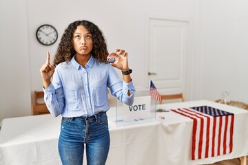 Beautiful hispanic woman standing by at political campaign by voting ballot pointing up looking sad and upset, indicating direction with fingers, unhappy and depressed.