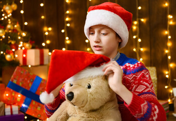 Child girl posing and having fun in new year or christmas decoration. Holiday lights and gifts, Christmas tree decorated with toys. She's wearing a red sweater and a Santa helper hat