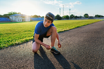 concept of sports and health - teen boy posing at a stadium track, he ties his shoelaces, a soccer field with green grass