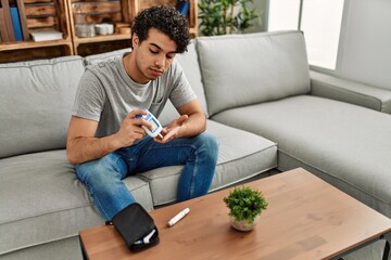 Young hispanic diabetic man measuring glucose sitting on the sofa at home.
