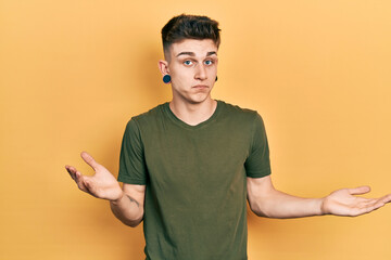 Young caucasian boy with ears dilation wearing casual green t shirt clueless and confused expression with arms and hands raised. doubt concept.