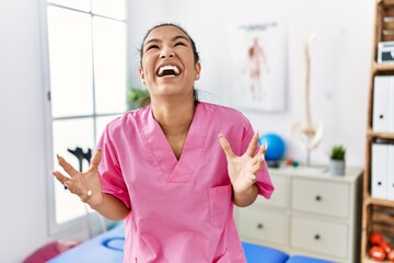 Young hispanic woman working at pain recovery clinic crazy and mad shouting and yelling with aggressive expression and arms raised. frustration concept.