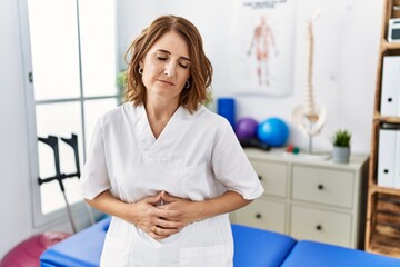 Middle age physiotherapist woman working at pain recovery clinic with hand on stomach because nausea, painful disease feeling unwell. ache concept.