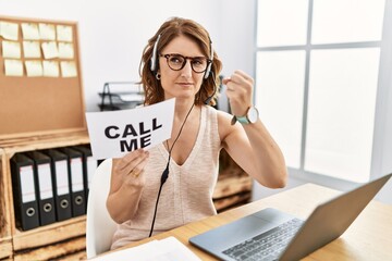 Middle age brunette woman wearing operator headset holding call me banner angry and mad raising...