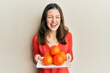 Young brunette woman holding plate with fresh oranges smiling and laughing hard out loud because funny crazy joke.