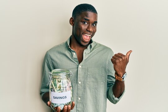Young african american man holding jar with savings pointing thumb up to the side smiling happy with open mouth