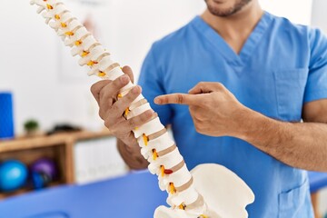 Young hispanic man wearing physio therapist uniform pointing to anatomical model of vertebral...