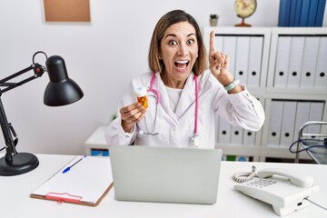 Young hispanic woman wearing doctor uniform holding pills at the clinic pointing finger up with successful idea. exited and happy. number one.