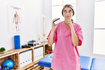 Young physiotherapist woman working at pain recovery clinic holding credit card serious face thinking about question with hand on chin, thoughtful about confusing idea