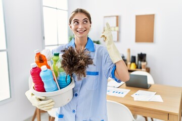 Young blonde woman wearing cleaner uniform holding cleaning products pointing finger up with successful idea. exited and happy. number one.