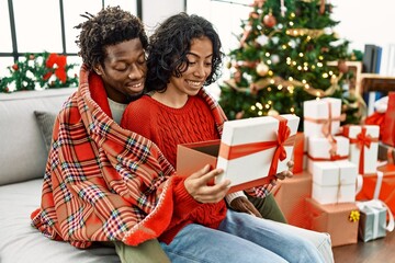 Young interracial couple smiling happy unboxing christmas gift sitting on the sofa at home.