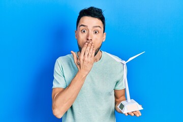 Young hispanic man holding solar windmill for renewable electricity covering mouth with hand,...