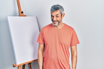 Handsome middle age man with grey hair standing by painter easel stand winking looking at the...