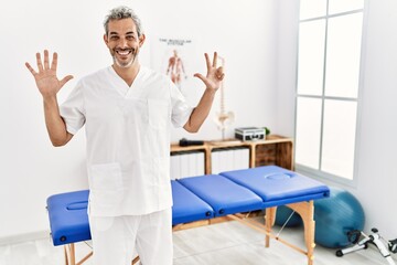 Middle age hispanic therapist man working at pain recovery clinic showing and pointing up with fingers number eight while smiling confident and happy.