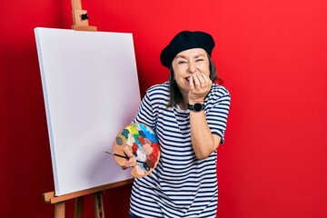 Middle age hispanic woman standing drawing with palette by painter easel stand laughing and...