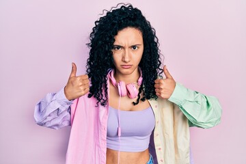 Young hispanic woman with curly hair doing thumbs up positive gesture skeptic and nervous, frowning upset because of problem. negative person.