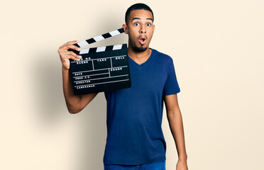 Young african american man holding video film clapboard scared and amazed with open mouth for...
