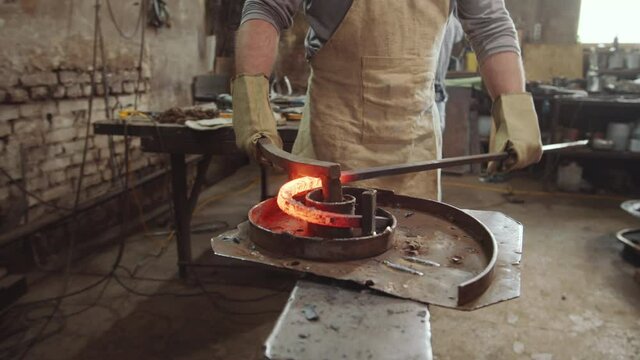 Midsection shot of blacksmith in apron and protective gloves shaping heated wrought iron rod into spiral in workshop