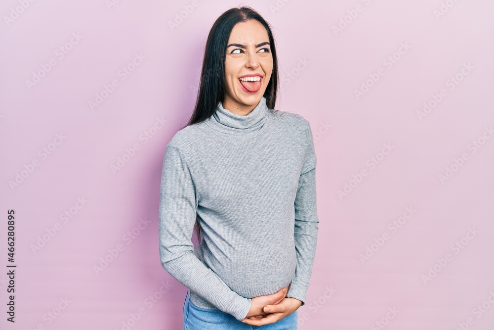 Sticker Beautiful woman with blue eyes expecting a baby, touching pregnant belly sticking tongue out happy with funny expression. emotion concept.