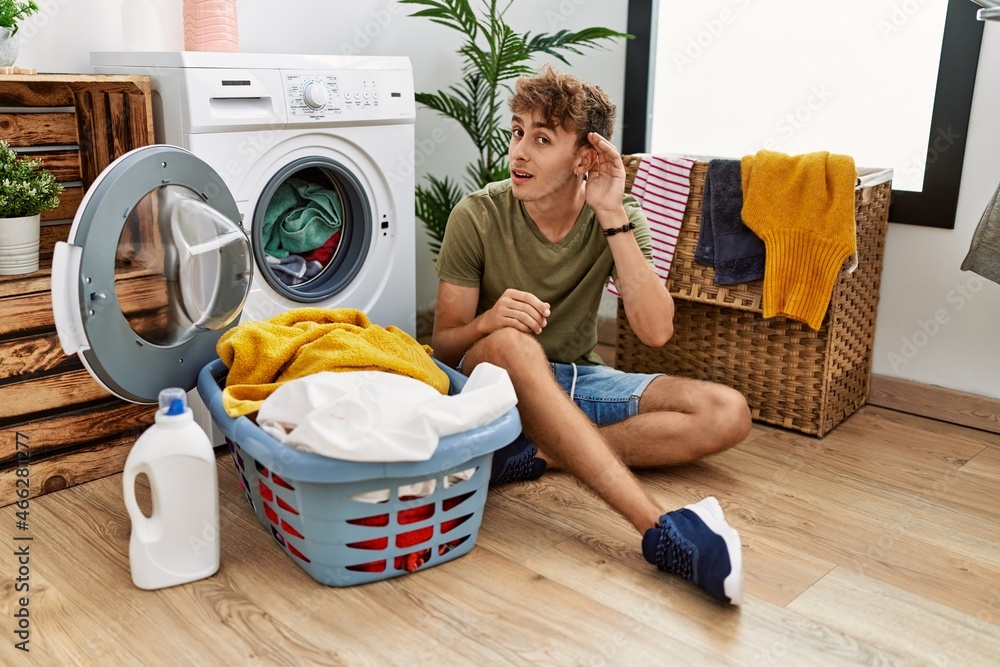 Sticker Young caucasian man putting dirty laundry into washing machine smiling with hand over ear listening an hearing to rumor or gossip. deafness concept.