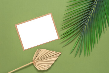 Blank invitation card mockup with dry palm leaf and artificial palm leaves. Green-olive paper background. Top view, flat lay.
