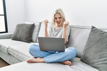 Young caucasian woman using laptop at home sitting on the sofa very happy and excited doing winner gesture with arms raised, smiling and screaming for success. celebration concept.